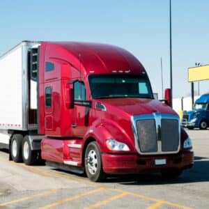 A red semi-truck with a white trailer, used for less-than-truckload shipping, is parked in a parking lot on a sunny day, with another blue truck visible in the background.