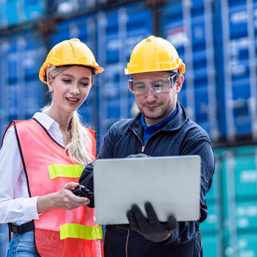 Two workers in safety gear and hard hats review data on a tablet in front of stacked shipping containers, ensuring seamless operations with Carrier Solutions for efficient logistics.