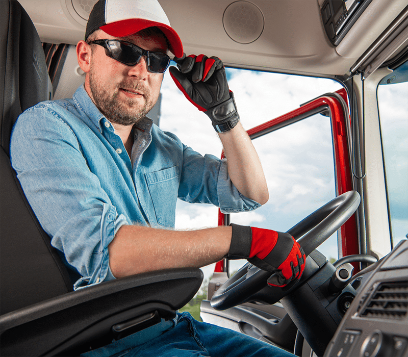 A man wearing a cap, sunglasses, and gloves sits in the driver's seat of a Carrier Solutions truck, gripping the steering wheel with one hand while adjusting his sunglasses with the other.