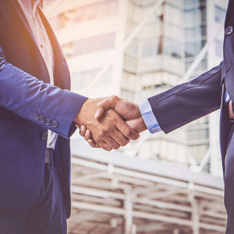 Two professionals in business attire shake hands confidently in front of a modern office building, sealing a strategic partnership with a third-party logistic company.