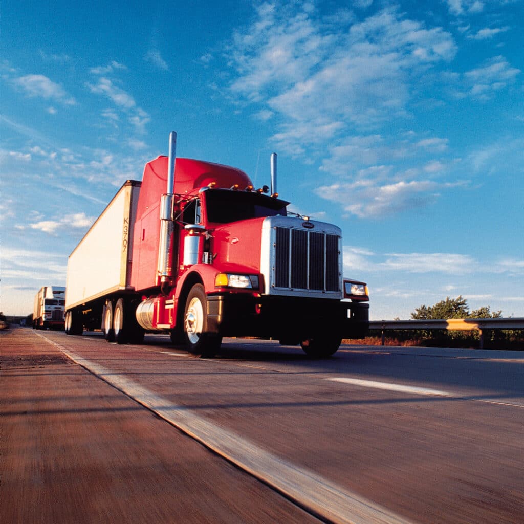 A red semi-truck from a third-party logistic company cruises down the highway beneath a clear blue sky dotted with scattered clouds.