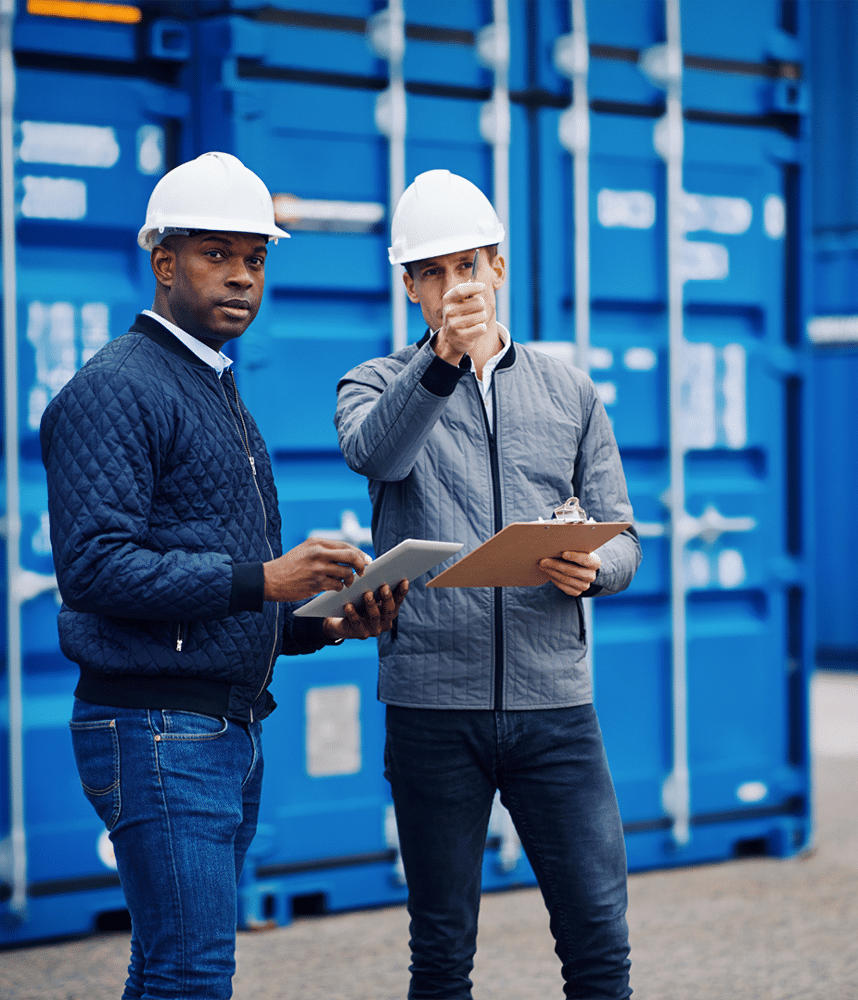 Two workers in hard hats stand in front of blue shipping containers; one holds a tablet showing advanced shipping software, while the other holds a clipboard and points forward, highlighting efficient supply chain management strategies.