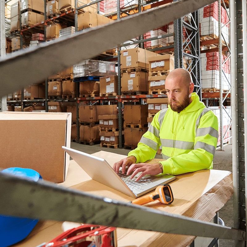 A person in a high-visibility jacket focuses on their laptop at a table, surrounded by boxes in the bustling warehouse. They're optimizing Carrier Solutions to enhance logistic efficiency.