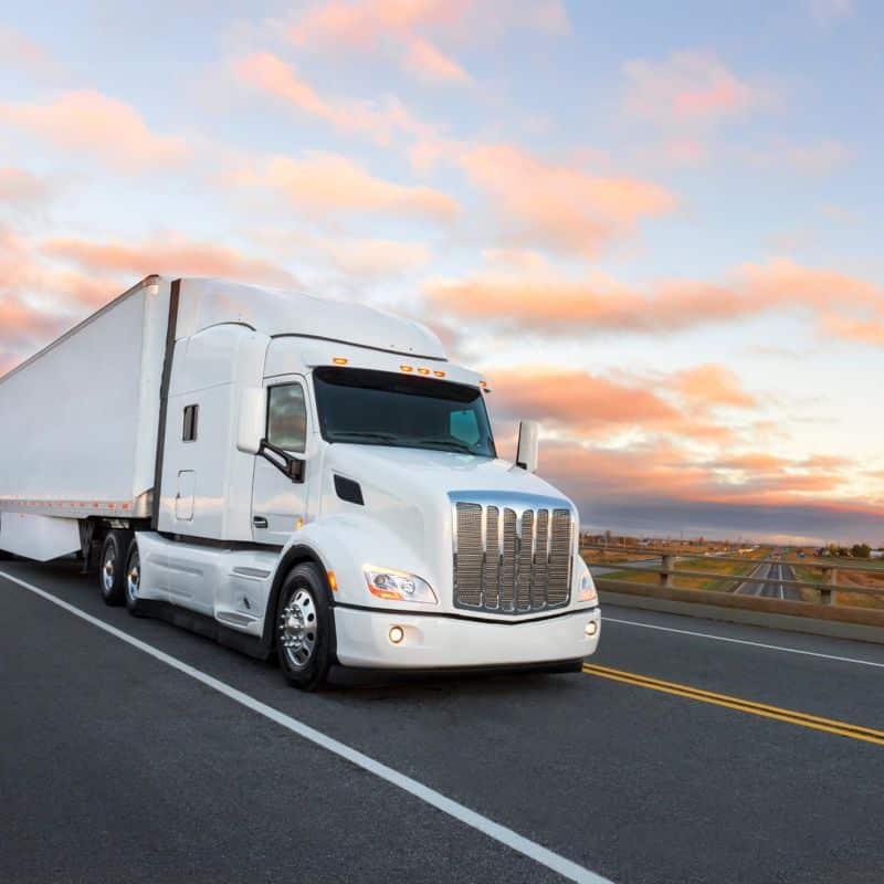 A white semi-truck with a trailer, representing a third-party logistic company, drives on a highway under a colorful sky at sunset.