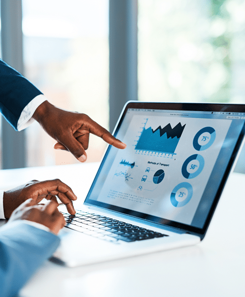 Two people at a desk examine financial graphs and charts on a laptop screen, exploring solutions. One person points at the screen, while the other types on the keyboard.