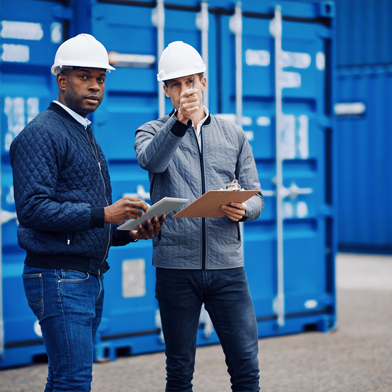 Two men in hard hats and jackets, one holding a clipboard and the other pointing, stand in front of blue shipping containers, discussing efficient solutions.