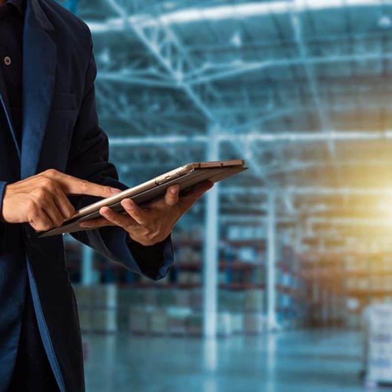 A person in a dark suit navigates a tablet within a spacious, well-lit warehouse. In the background, the shelves blur softly—a busy hub for a third-party logistic company ensuring seamless operations.