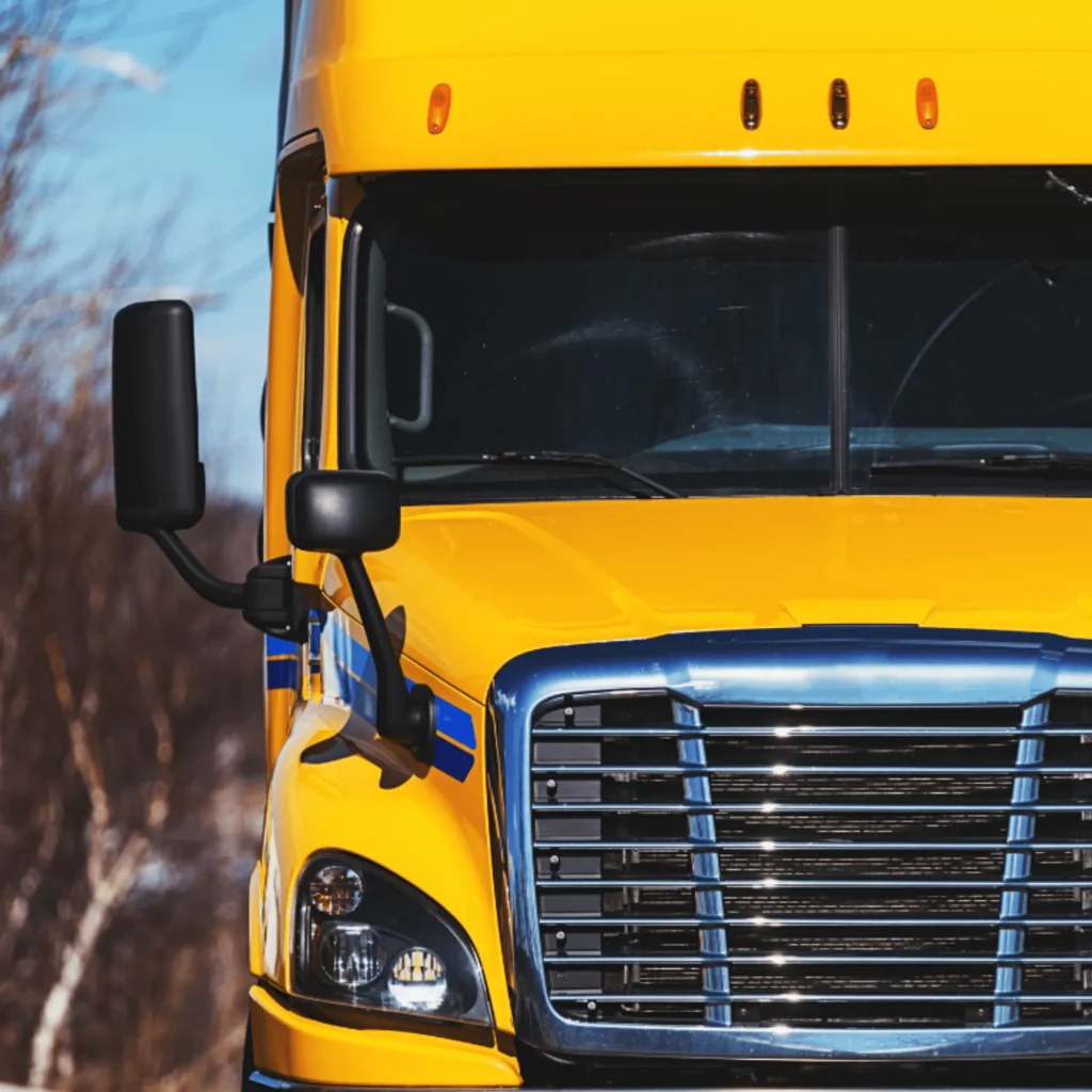 Front view of a yellow semi-truck with a chrome grille and large side mirrors, symbolizing the backbone of logistics, set against a blurred outdoor background.