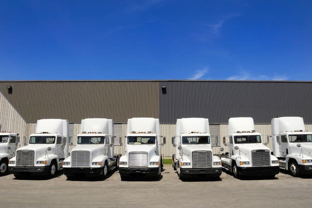 A row of white semi-trucks, vital for Carrier Management, is parked in front of a large industrial warehouse under a clear blue sky.