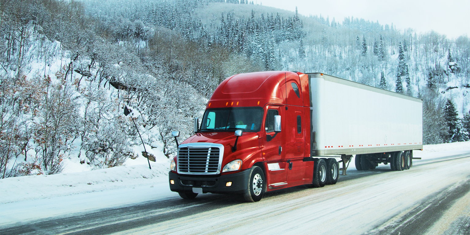 A red semi-truck, expertly managed through real-time tracking, drives on a snow-covered road through a hilly, tree-lined landscape in winter.