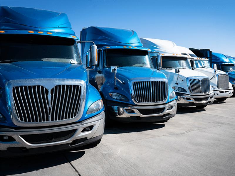 A row of blue and white semi-trucks, representing premier carrier solutions, stands proudly on a concrete lot beneath a clear blue sky.