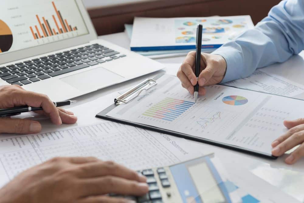 Two people review financial charts and graphs on paper with a laptop, calculator, and documents on a desk, analyzing business data and Carrier Management statistics.