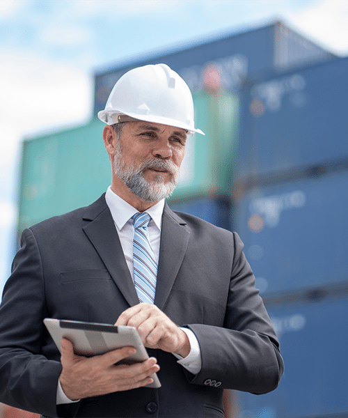 A man in a suit and hard hat stands before stacked shipping containers, expertly using a tablet to streamline Carrier Solutions tasks.