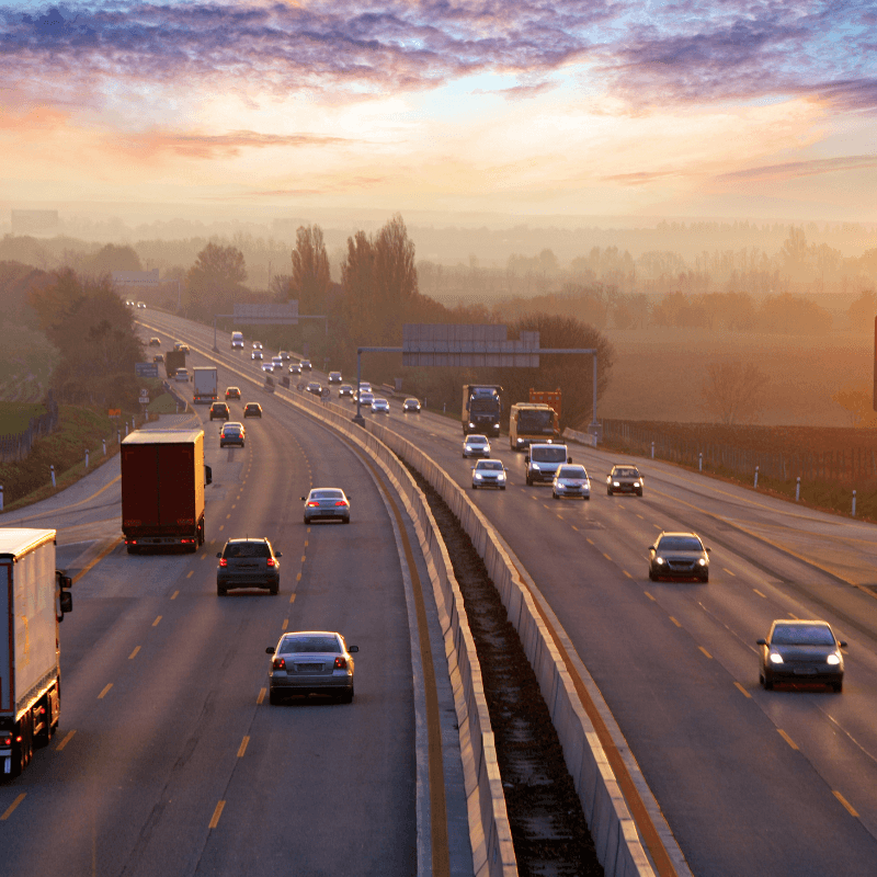 A multi-lane highway at sunset buzzes with cars and trucks, including fleets from third-party logistic companies, traveling in both directions. The sky is partly cloudy, casting a gentle glow over the rural landscape.