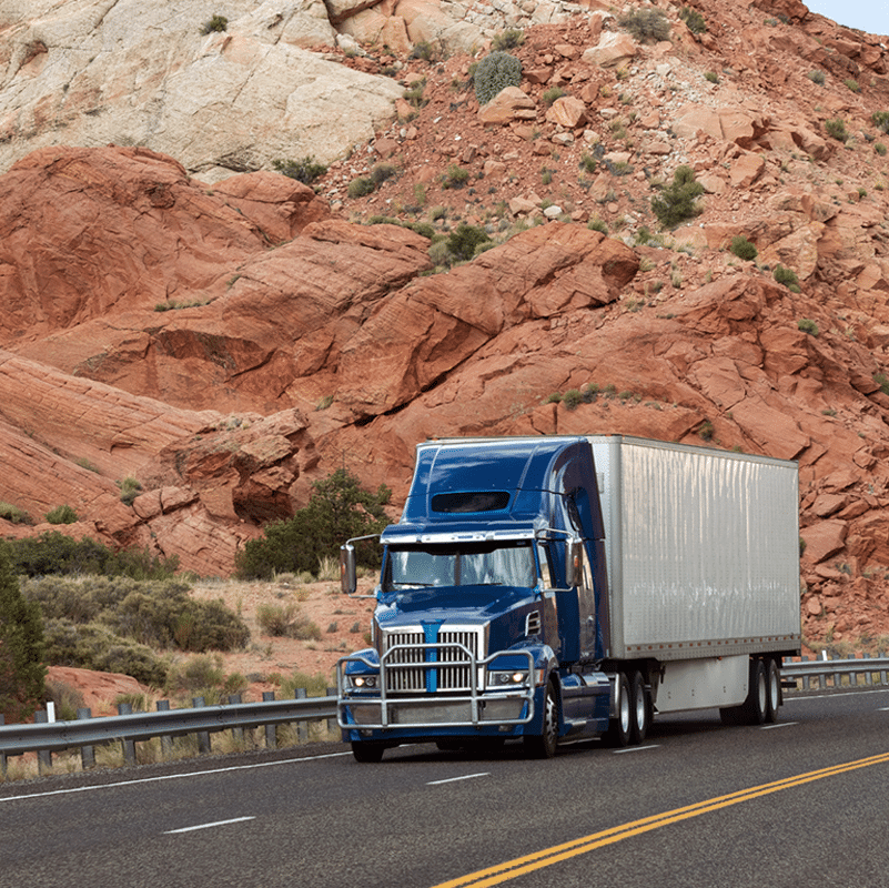 Blue semi-truck with a silver trailer, delivering shipping solutions as it drives on a highway through a rocky desert landscape.