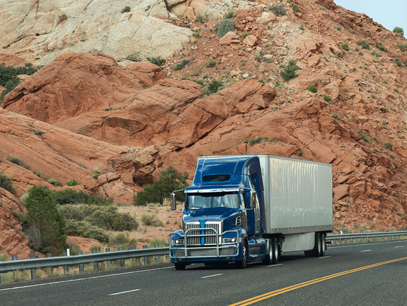 A blue semi-truck, representing innovative shipping solutions, travels on a highway with red rock formations in the background.