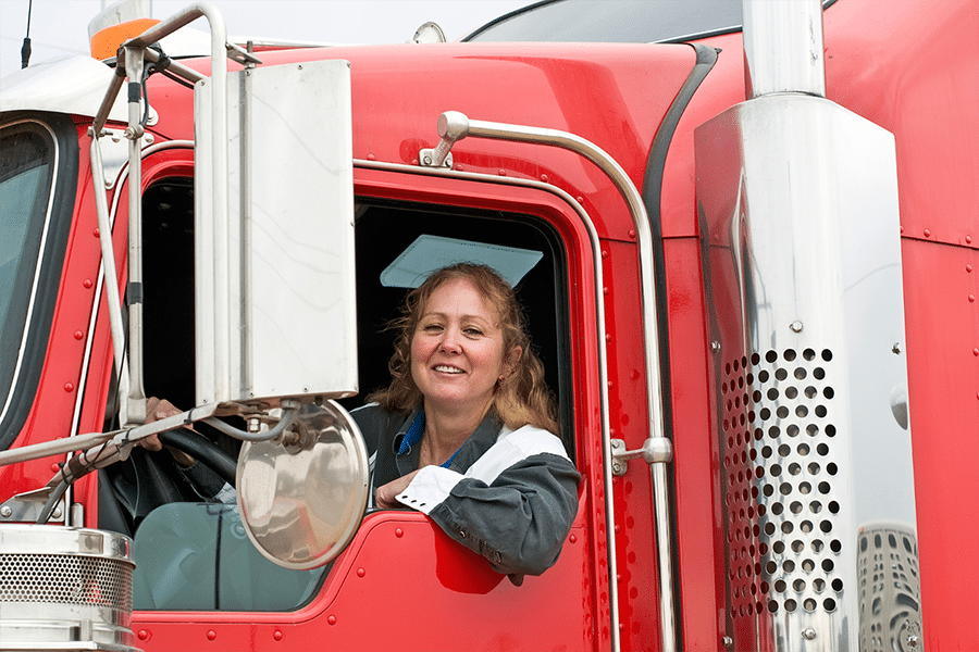 A person smiles brightly from the driver's seat of a red semi-truck, the window rolled down, proudly representing their third-party logistic company.